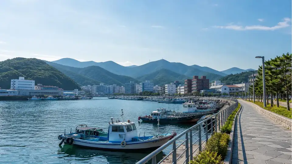 Wide summer view of a calm Korean harbor with green hills, a promenade and small fishing boats