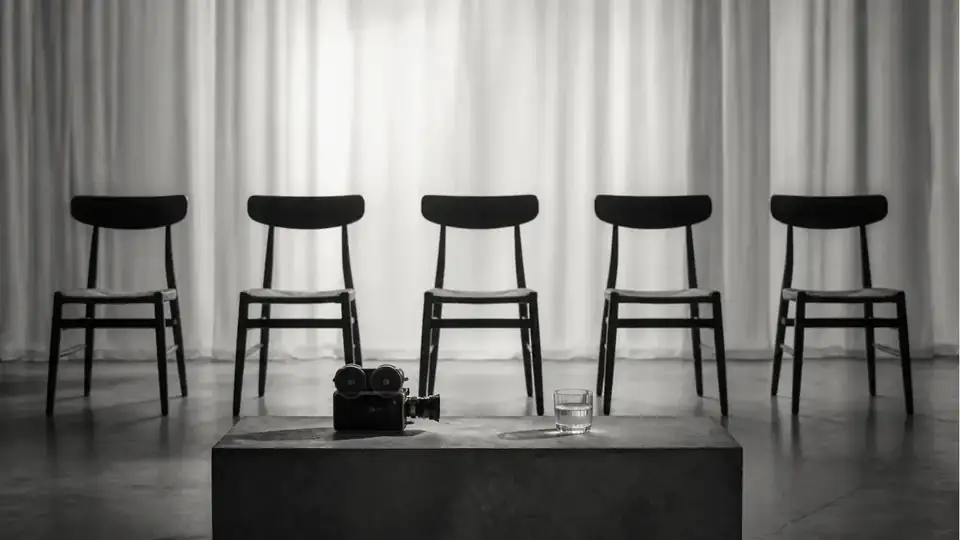 Minimal black-and-white studio scene with five empty chairs, soft curtains and a film camera